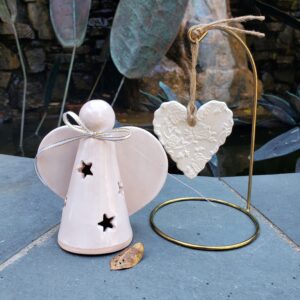 Handcrafted clay angel with stars cut out of the body for light to shine through, a white and gold ribbon is tied in a bow around the neck. Next to it is the ornament hanging on a golden stand by twine. The ornament is heart shaped with a rose pattern covering it. Both items are covered in a white glaze.
