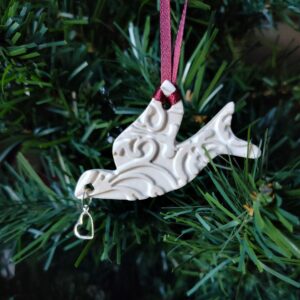 White dove ornament with a heart charm attached to its mouth. A red ribbon is used to hang the ornament from a tree branch.